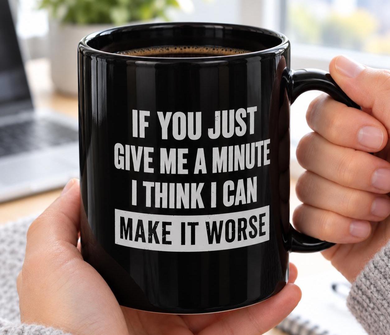 Hands holding the black Give Me A Minute I Can Make It Worse mug at a desk