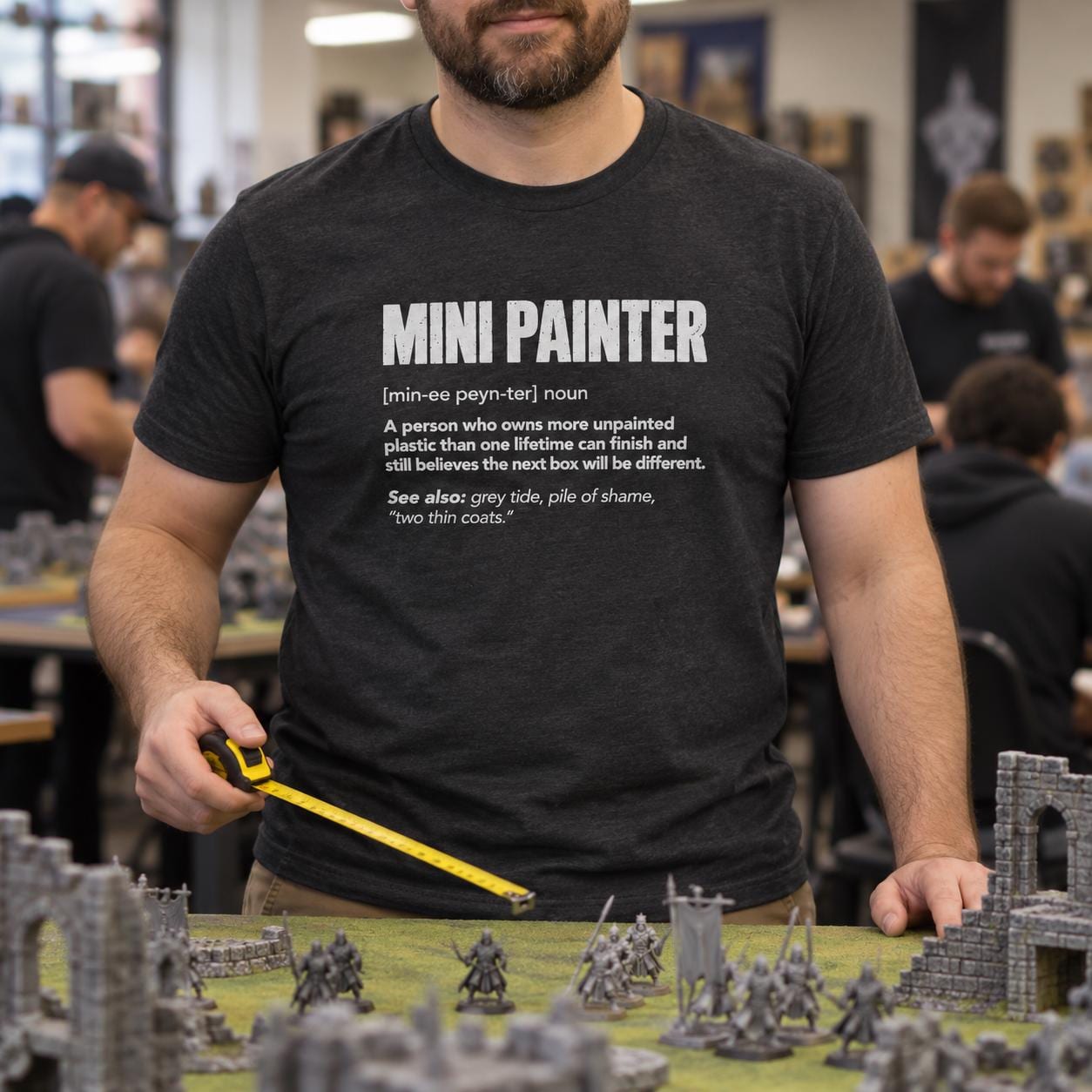Mini Painter Definition T-Shirt - man in the dark heather t-shirt measuring on a wargaming table