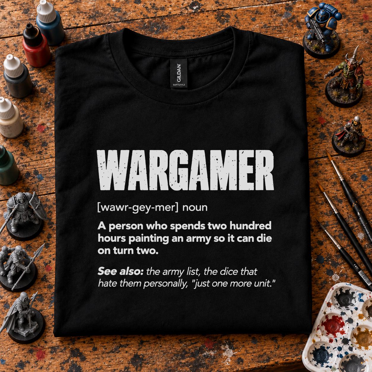 Wargamer Definition T-Shirt - black t-shirt folded on a paint splattered hobby desk with miniatures, paint bottles and brushes