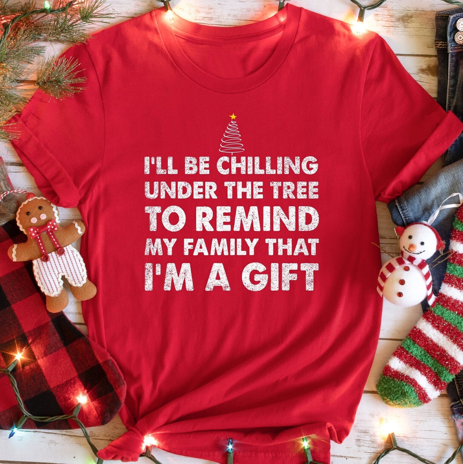 A red t-shirt with the text I'LL BE CHILLING UNDER THE TREE TO REMIND MY FAMILY THAT I'M A GIFT and a Christmas tree graphic, surrounded by Christmas decorations.