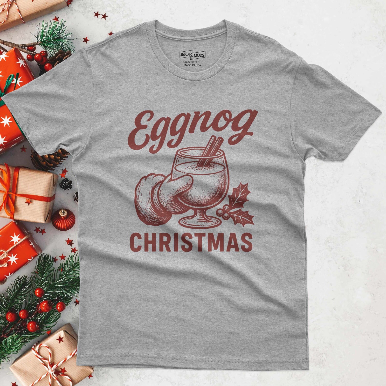 A heather gray t-shirt with a festive design. The graphic features the word "Eggnog" in a stylized font, along with an illustration of an eggnog drink in a glass, a nutmeg, and the word "Christmas".