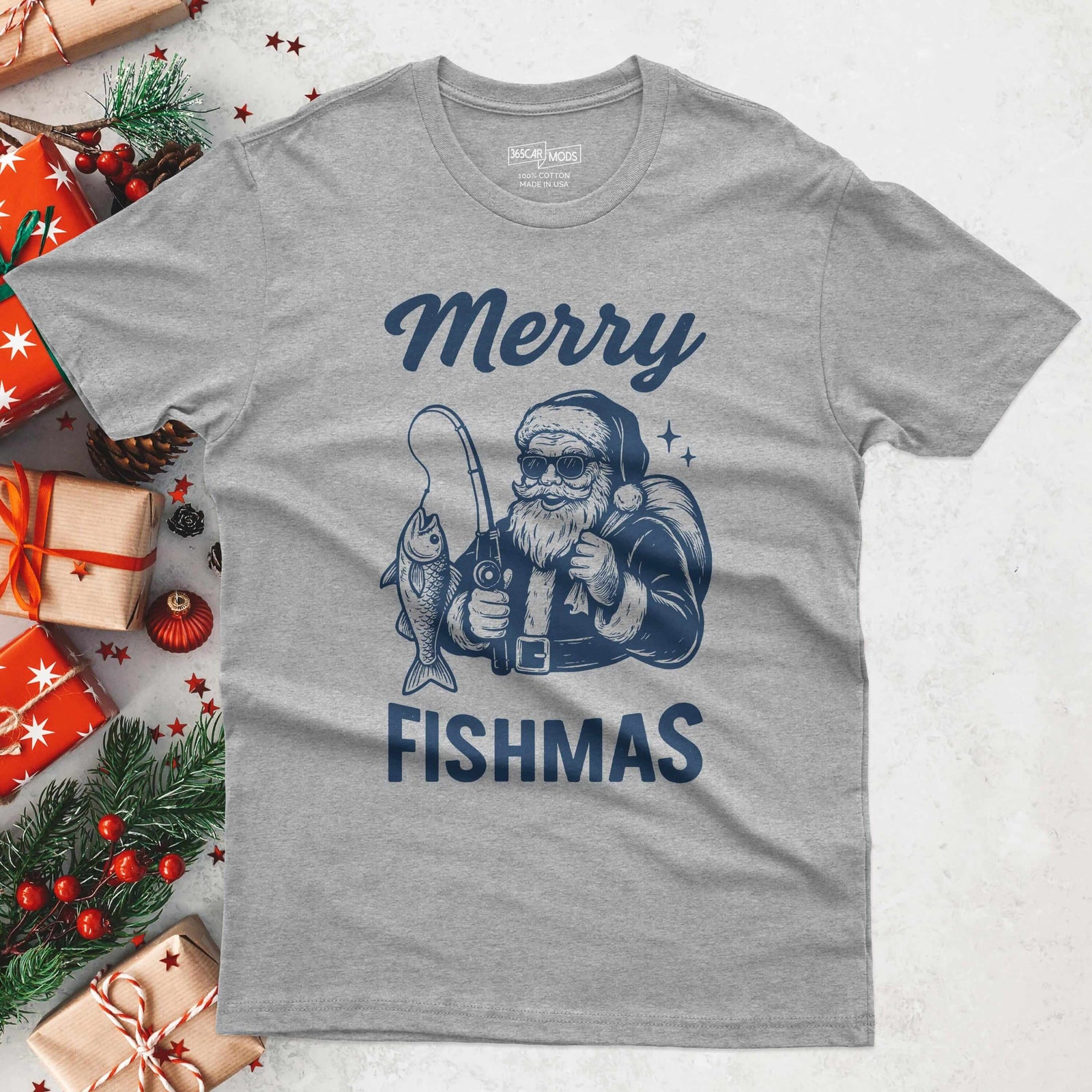 A gray t-shirt with a graphic of Santa Claus fishing, holding a fishing rod with a fish on the line. The text "Merry Fishmas" is printed in navy blue above and below the graphic.