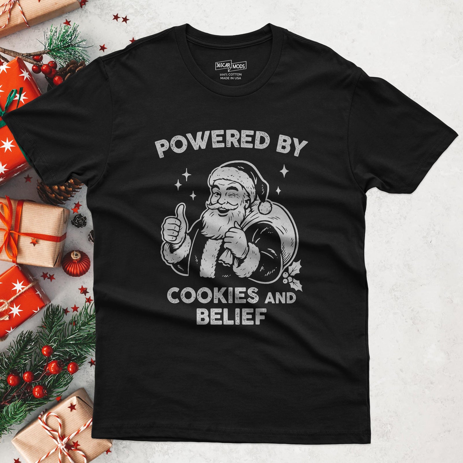 Black t-shirt with a graphic of Santa Claus giving a thumbs up next to a sack of gifts and the text POWERED BY COOKIES AND BELIEF.