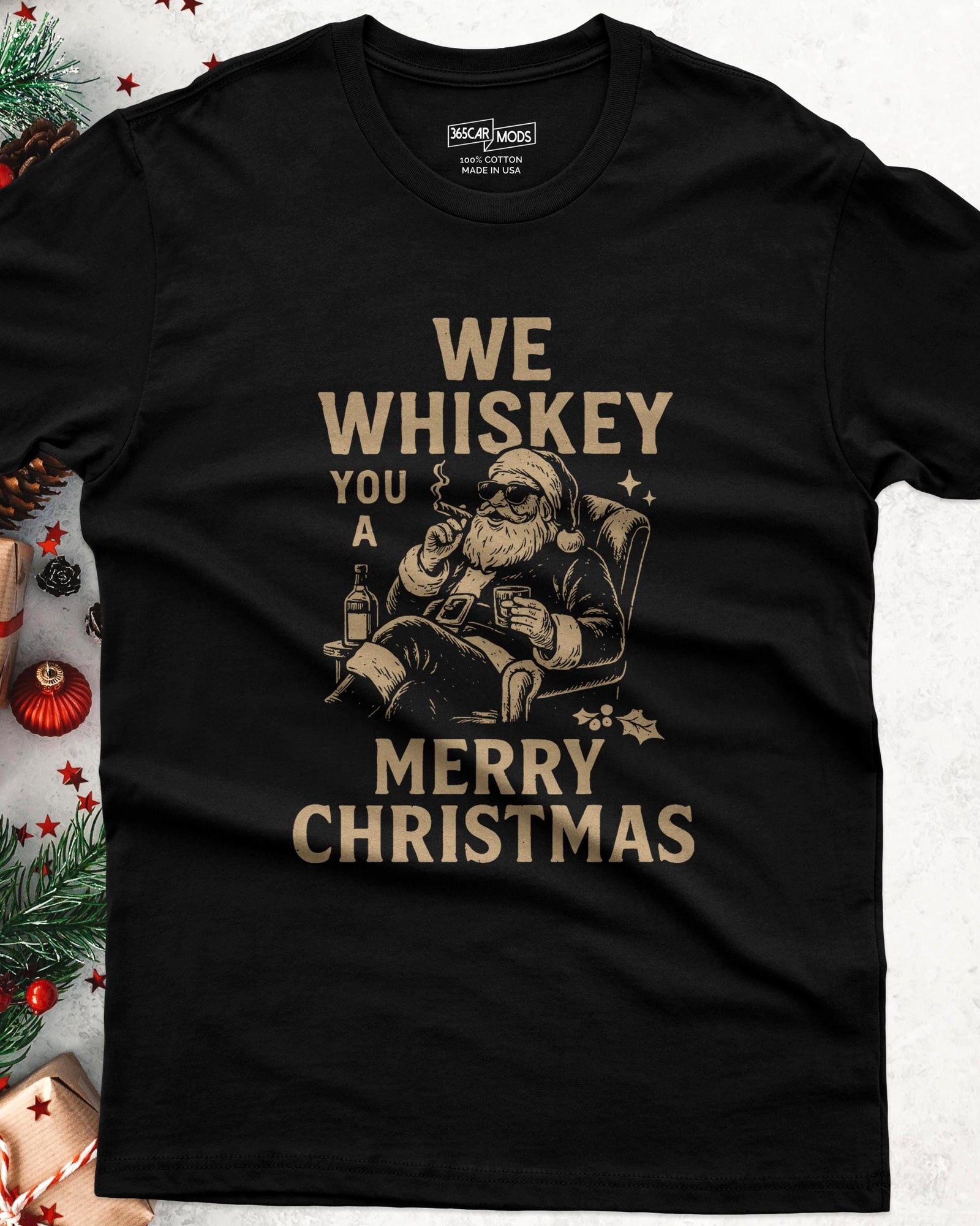 A black short-sleeve t-shirt featuring a graphic of Santa Claus wearing sunglasses, sitting in a chair, holding a glass and a cigar. The text reads: WE WHISKEY YOU A MERRY CHRISTMAS.