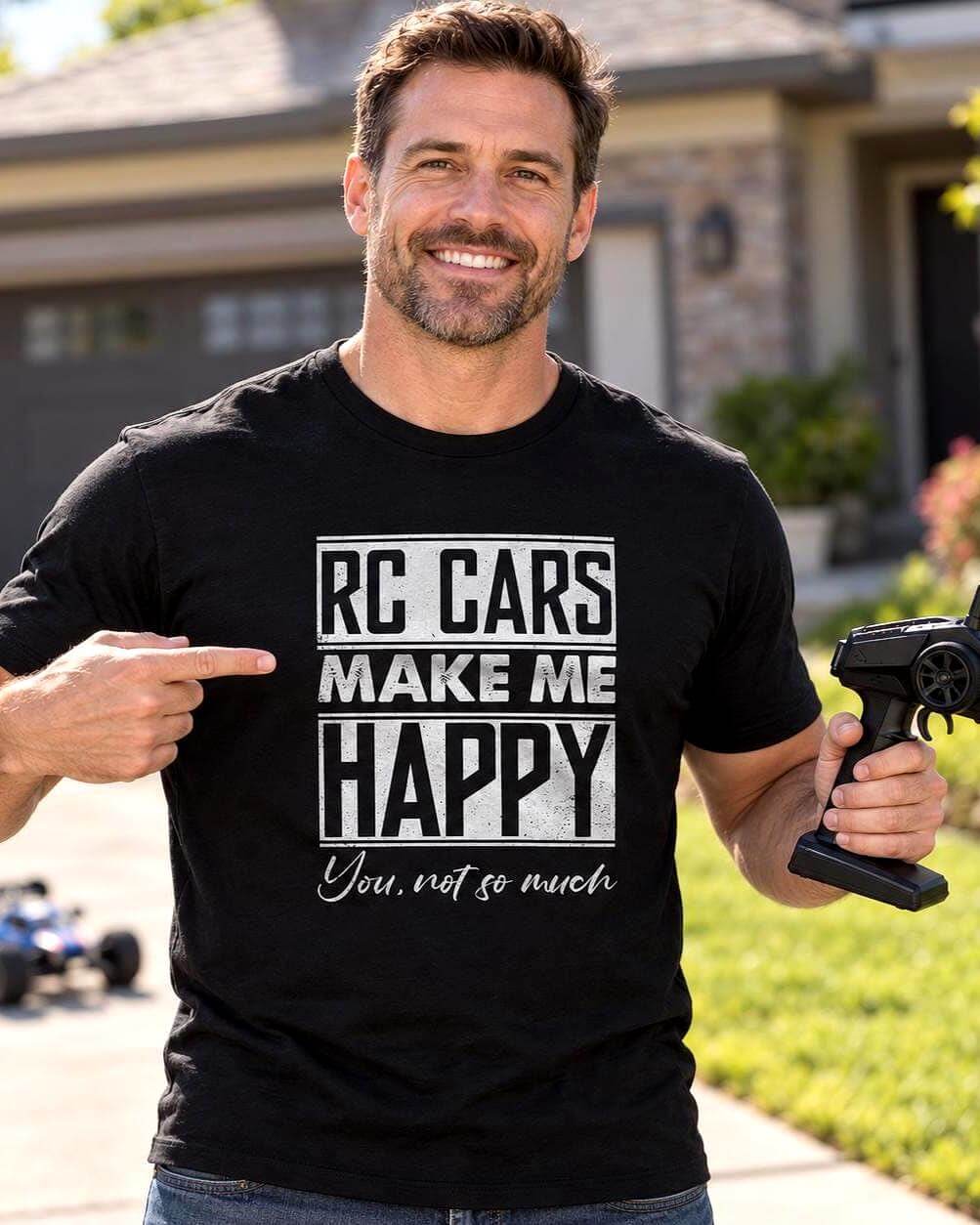 Smiling man wearing a black “RC Cars Make Me Happy, You Not So Much” T-shirt, holding an RC car controller and pointing to the design outside his suburban home.
