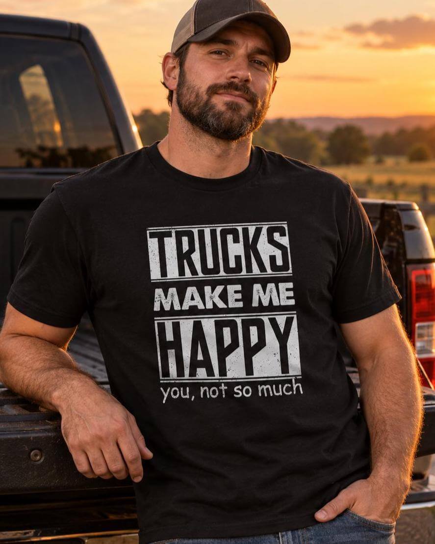 Man in a black funny truck lover shirt leaning on the tailgate of a lifted pickup truck on a gravel driveway at sunset