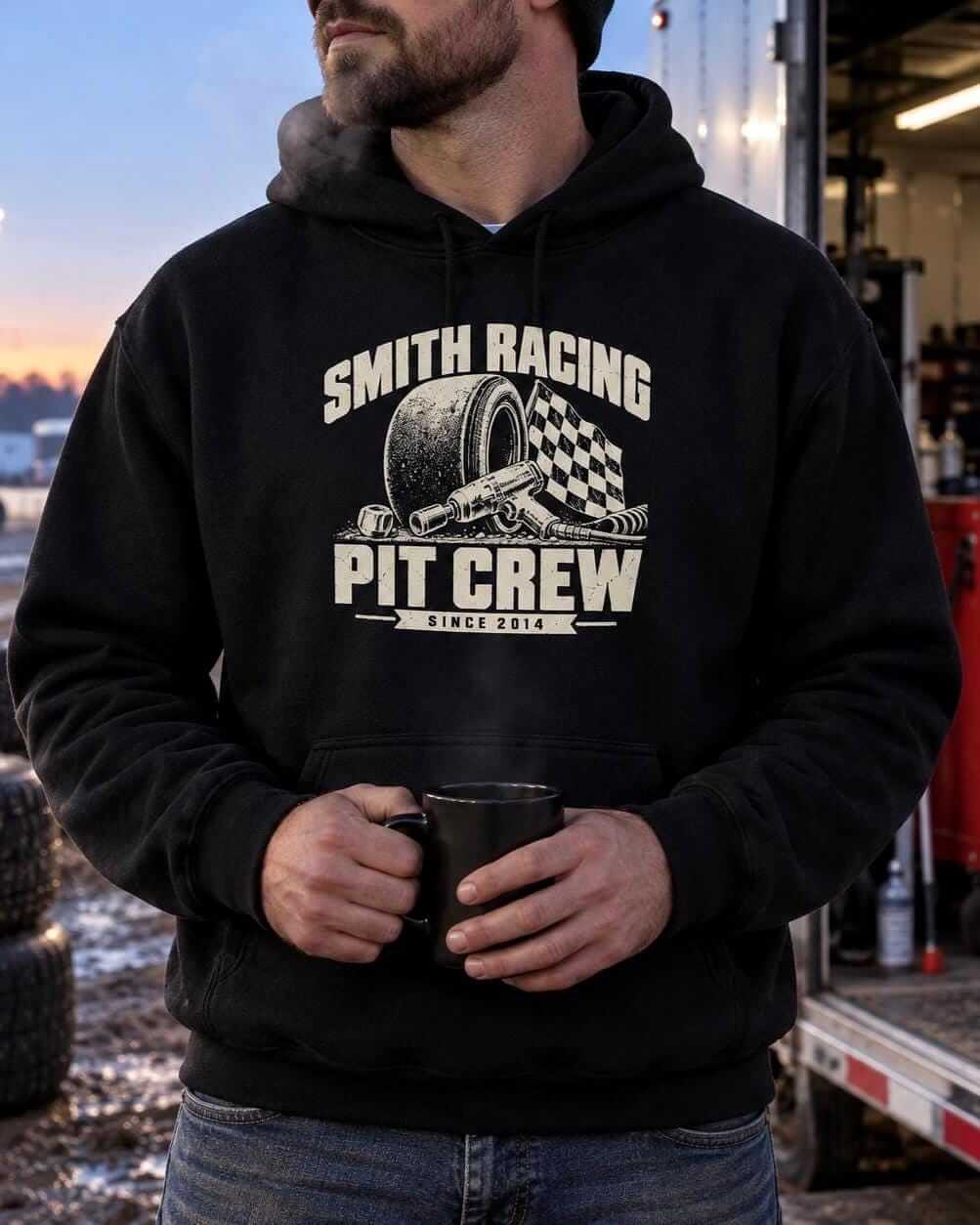 Man wearing the black personalized Pit Crew racing hoodie and holding a coffee mug next to a race trailer at dusk