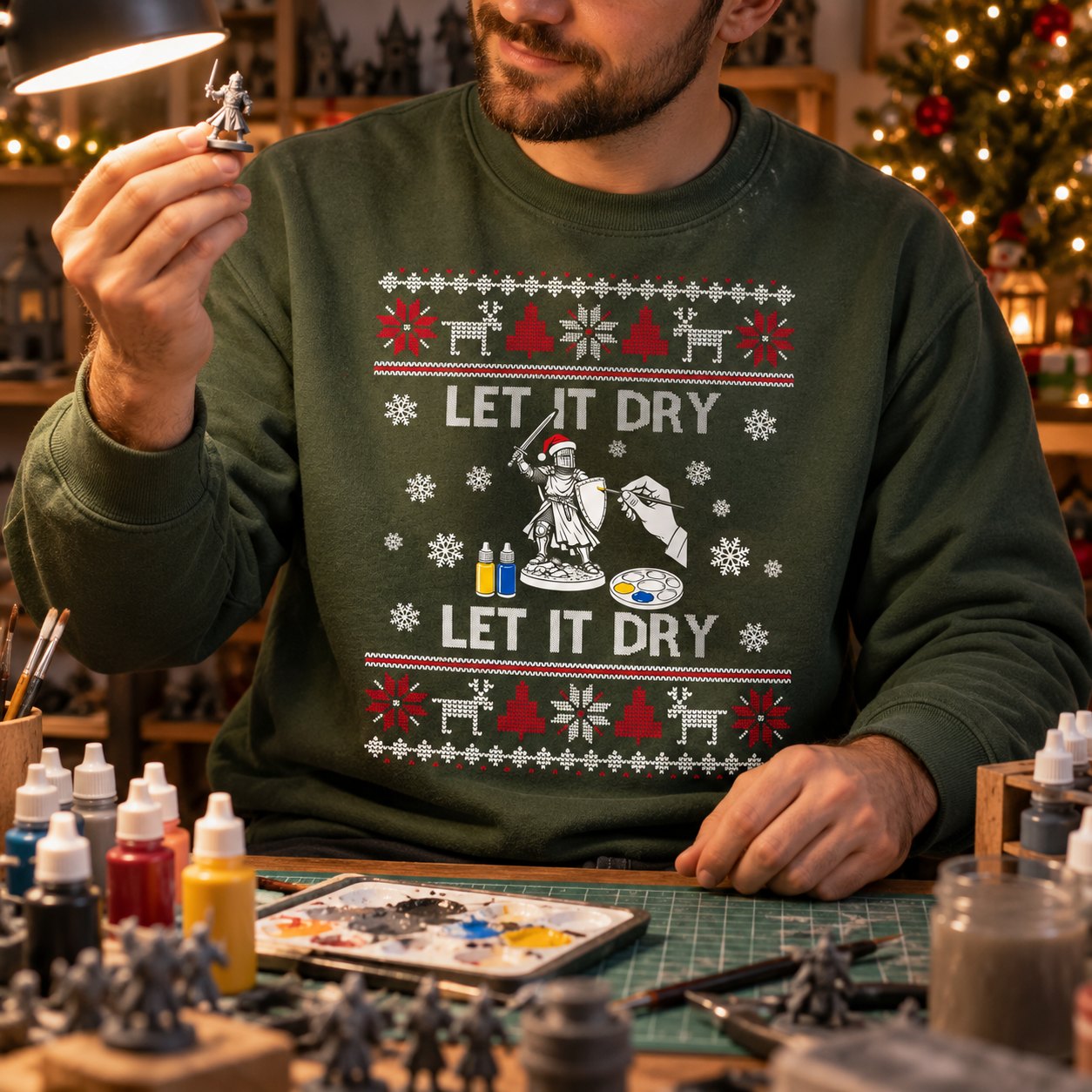 Miniature painter in the military green Let It Dry Christmas sweatshirt holding up a painted knight at his hobby desk