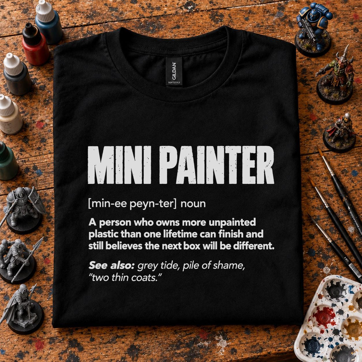 Mini Painter Definition T-Shirt - black t-shirt folded on a paint splattered hobby desk with miniatures, paint bottles and brushes