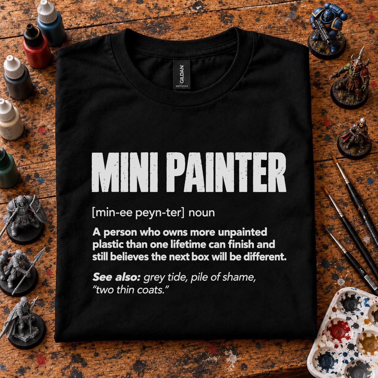 Mini Painter Definition T-Shirt - black t-shirt folded on a paint splattered hobby desk with miniatures, paint bottles and brushes