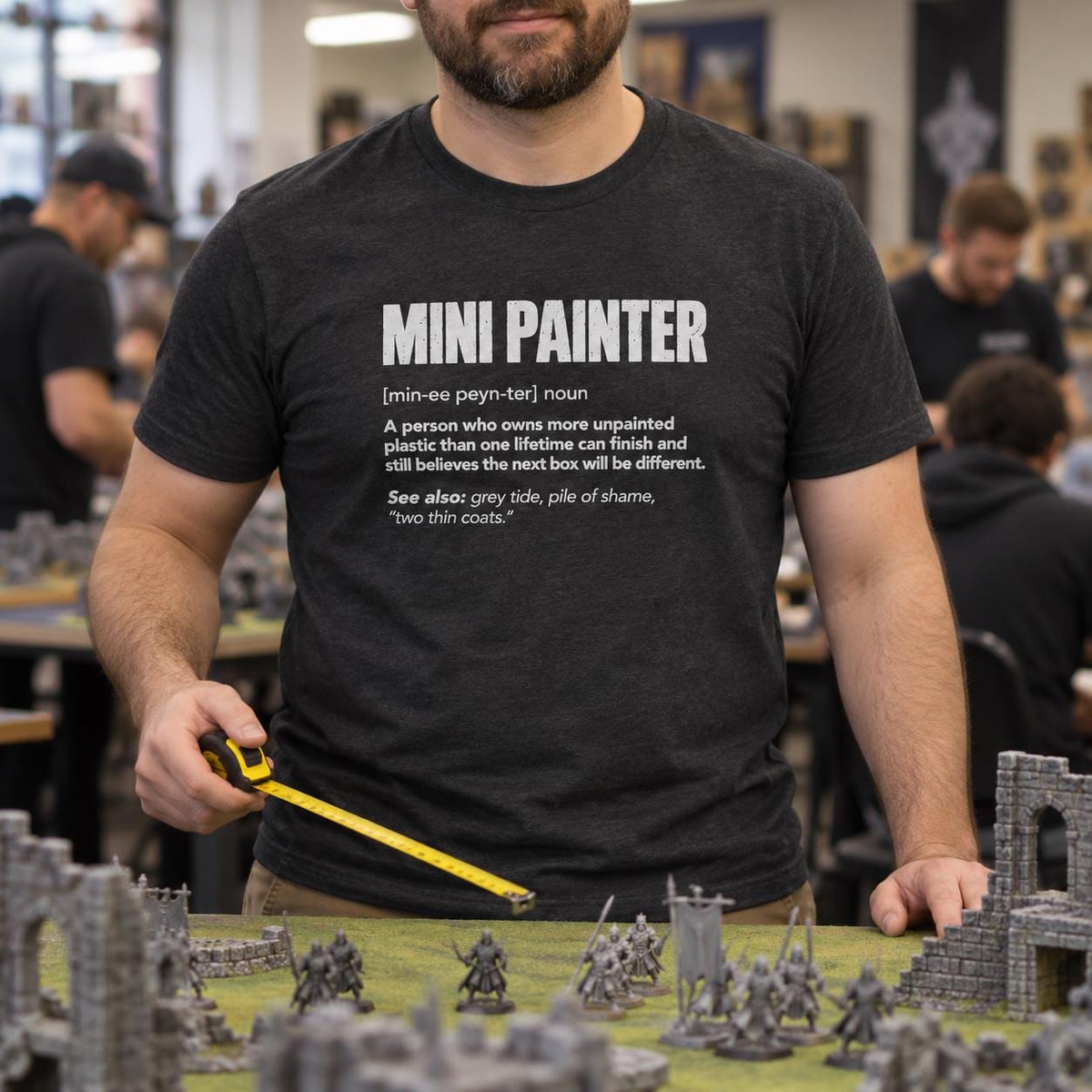 Mini Painter Definition T-Shirt - man in the dark heather t-shirt measuring on a wargaming table