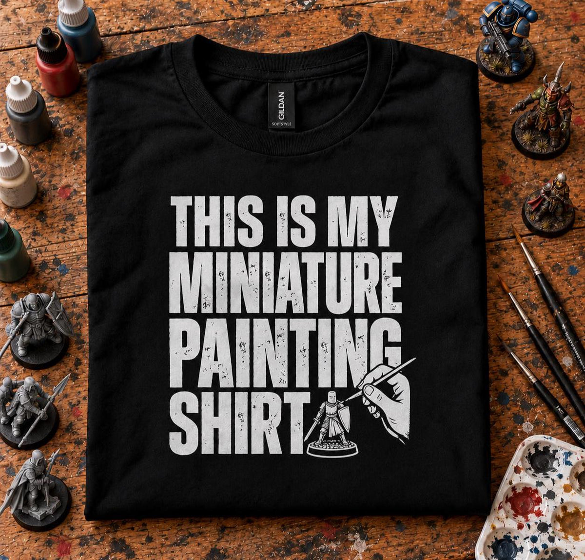 This Is My Miniature Painting Shirt - black t-shirt folded on a hobby desk with paints, brushes, a palette and miniatures