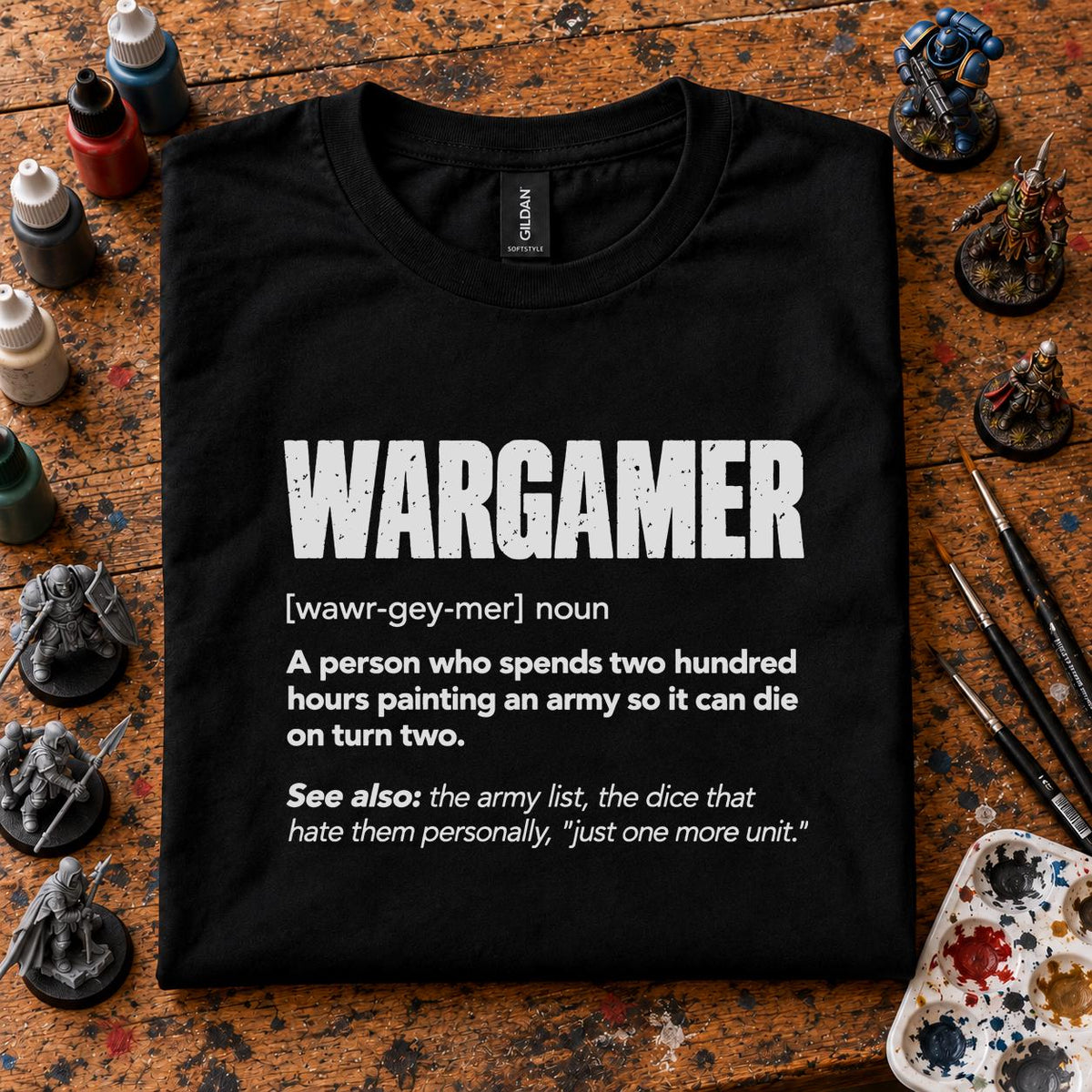 Wargamer Definition T-Shirt - black t-shirt folded on a paint splattered hobby desk with miniatures, paint bottles and brushes