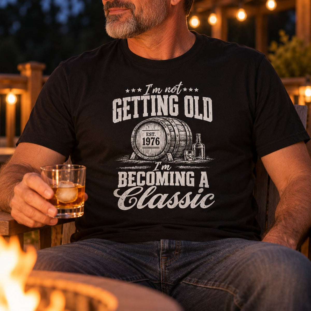 Man in the black Becoming A Classic whiskey t-shirt holding a glass of whiskey by a fire pit