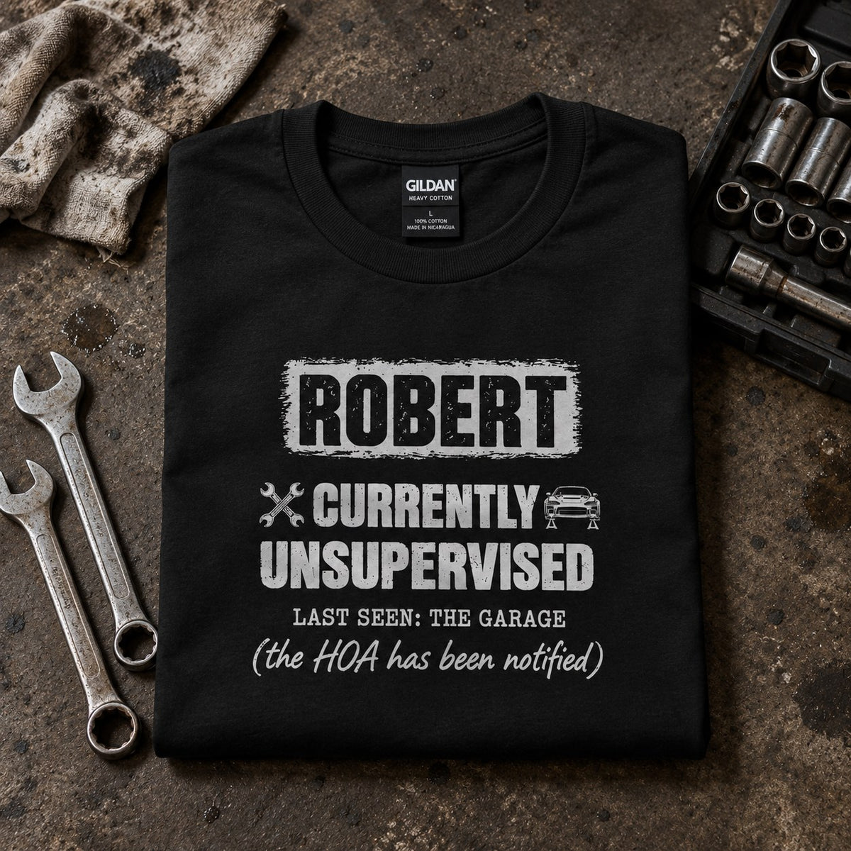 Black personalized Currently Unsupervised car guy t-shirt folded on a workbench next to wrenches and sockets