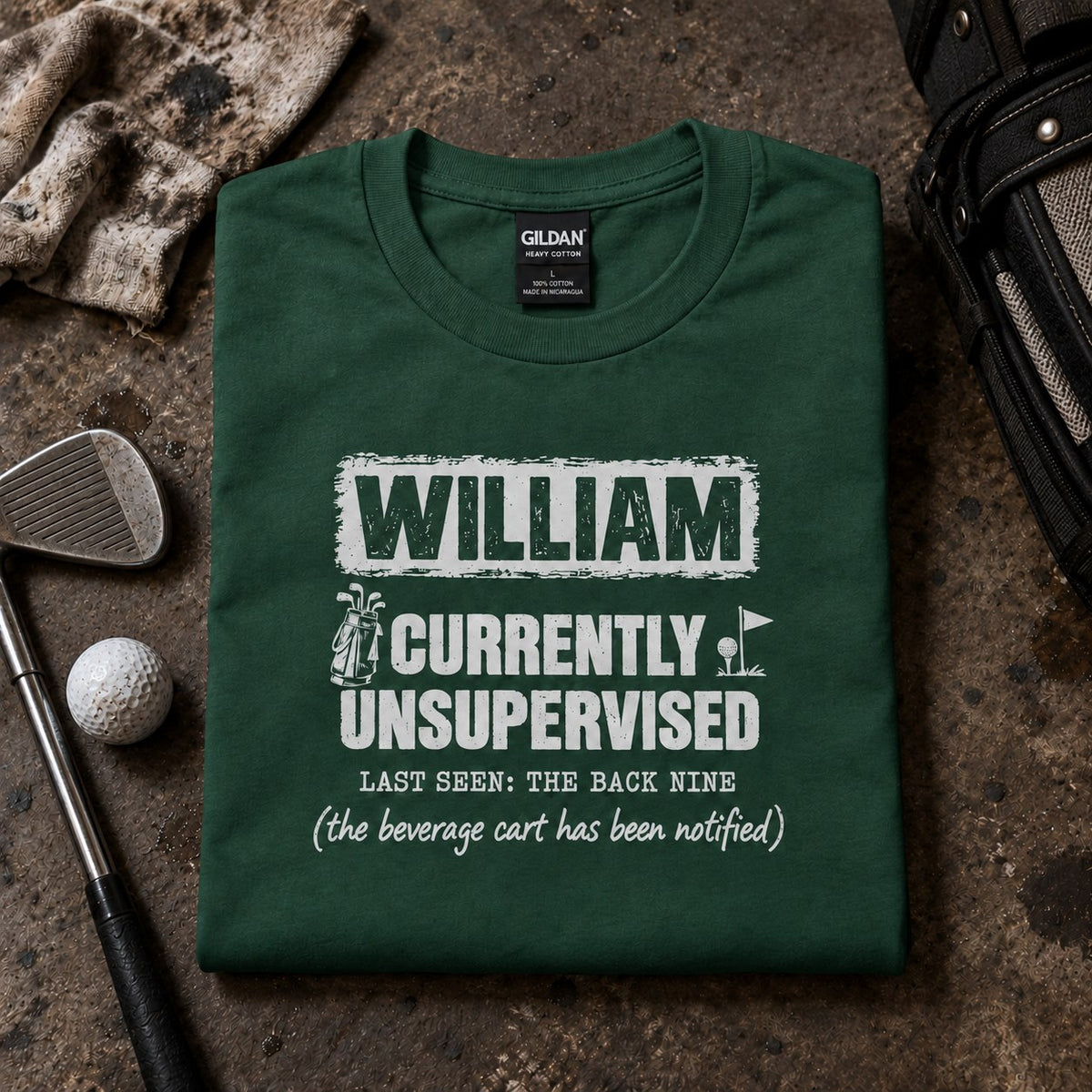 Forest green personalized Currently Unsupervised golf t-shirt folded next to golf balls and an iron