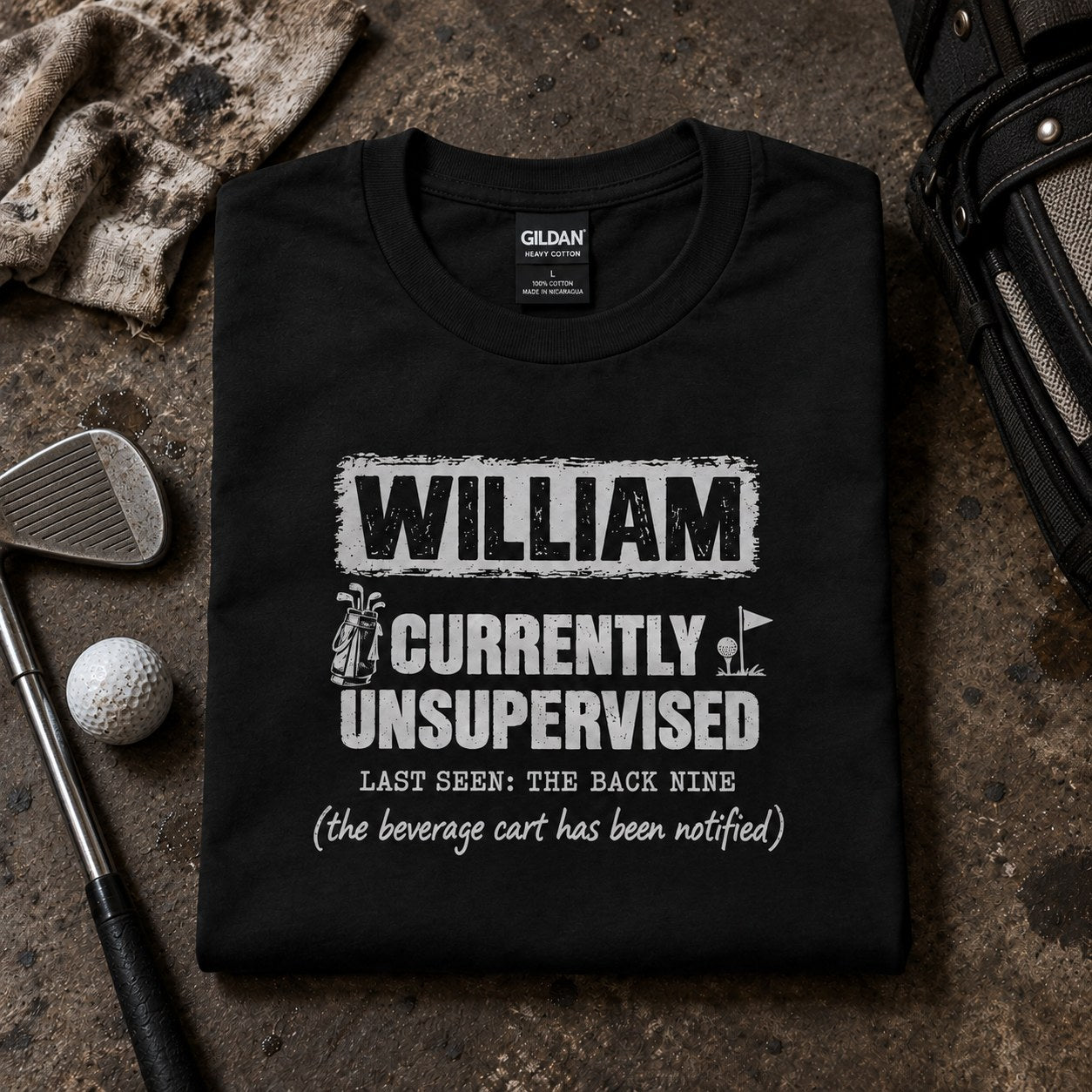 Black personalized Currently Unsupervised golf t-shirt folded next to golf balls and an iron