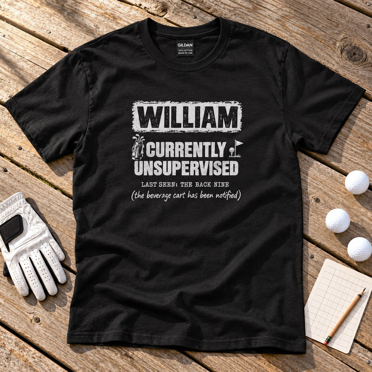 Black personalized Currently Unsupervised golf t-shirt on a wooden deck with golf balls and a glove