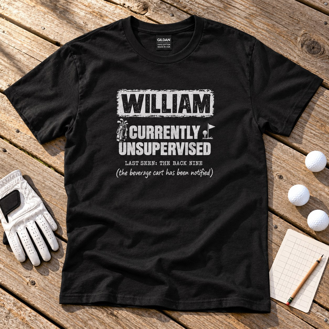 Black personalized Currently Unsupervised golf t-shirt on a wooden deck with golf balls and a glove