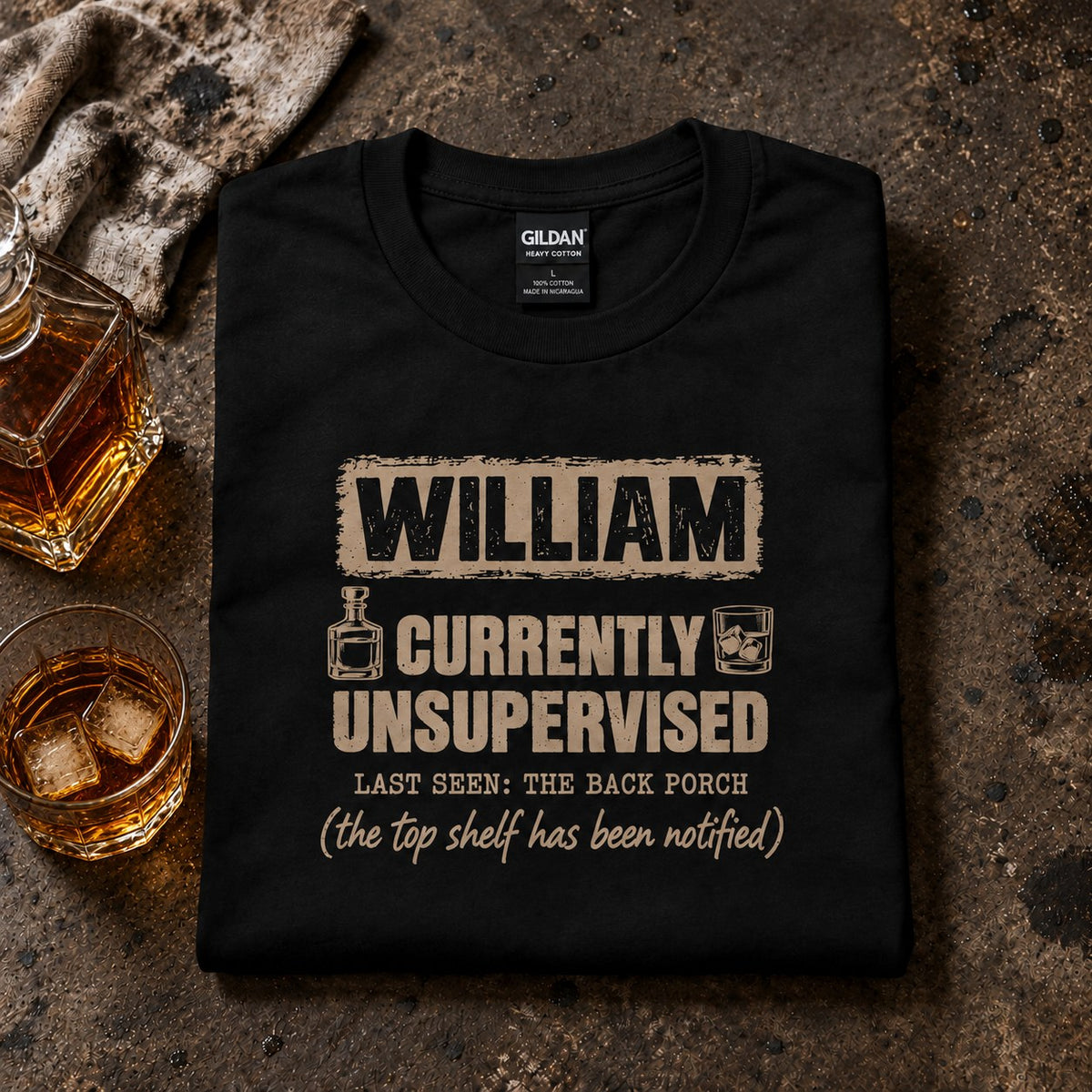 Black personalized Currently Unsupervised whiskey t-shirt folded next to a decanter and a glass of whiskey