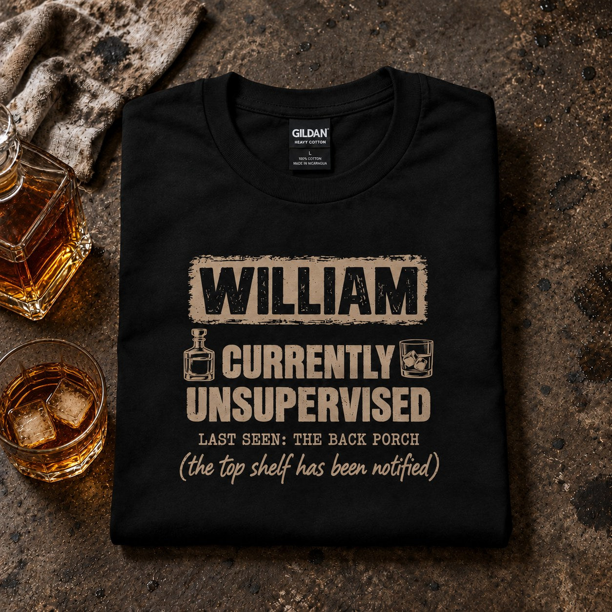 Black personalized Currently Unsupervised whiskey t-shirt folded next to a decanter and a glass of whiskey