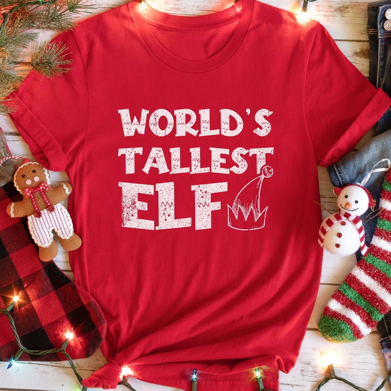 A red t-shirt with the text WORLD'S TALLEST ELF in white and an elf hat illustration, with festive decorations in the background.