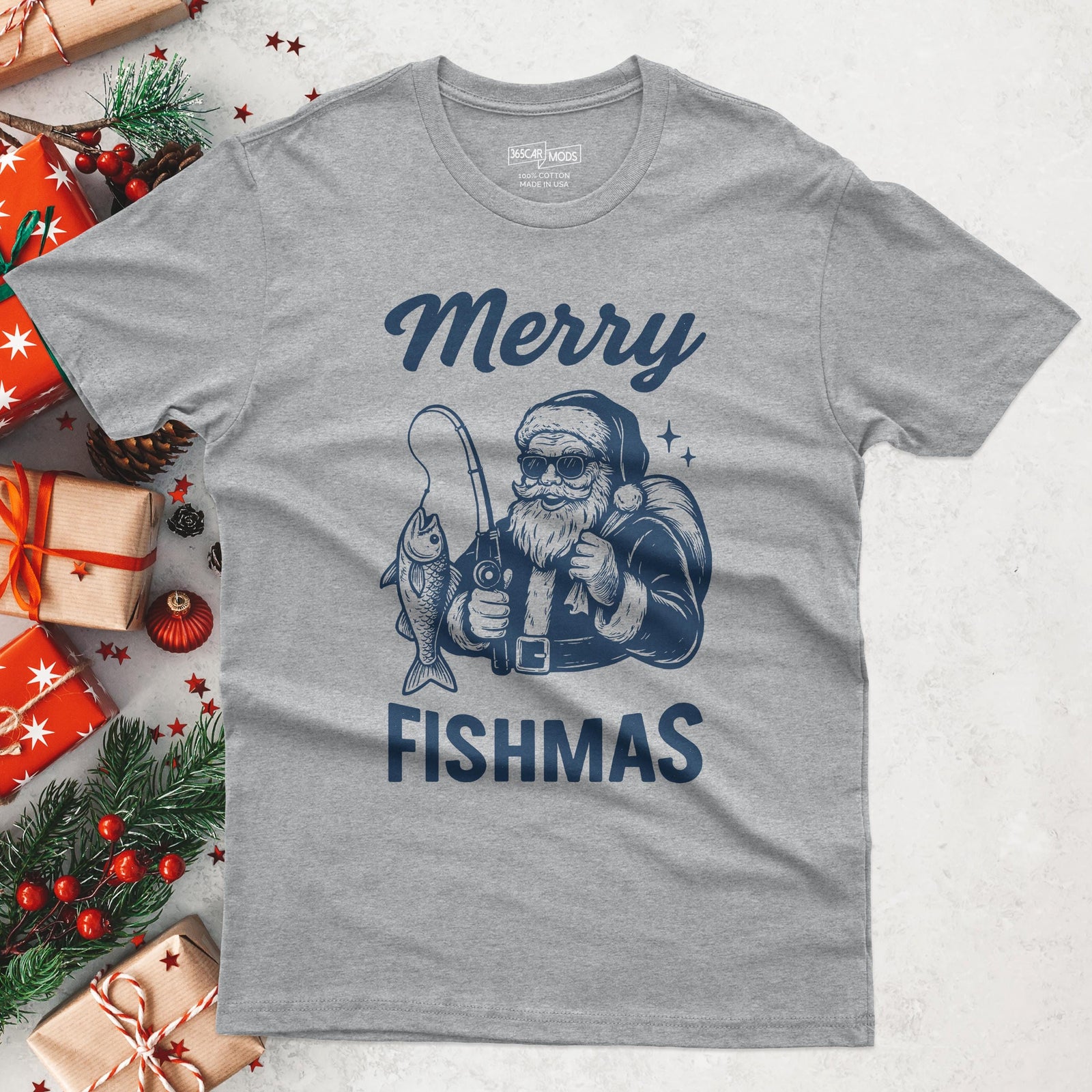 A gray t-shirt with a graphic of Santa Claus fishing, holding a fishing rod with a fish on the line. The text "Merry Fishmas" is printed in navy blue above and below the graphic.