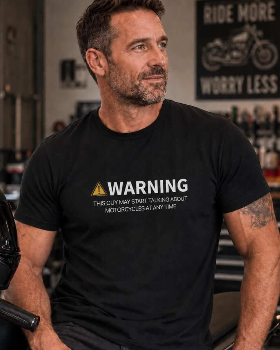 Man wearing a black T-shirt with a yellow warning symbol and the words “Warning, this guy may start talking about motorcycles at any time,” seated beside a motorcycle in a garage.