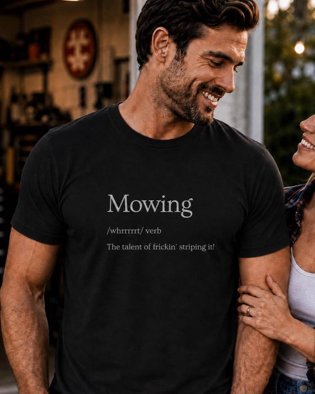 Man wearing a black Mowing Definition T-shirt beside a lawn mower on a freshly striped green lawn.