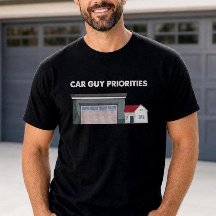 Smiling man wearing a black Car Guy Priorities graphic T-shirt while standing in front of a modern home garage.