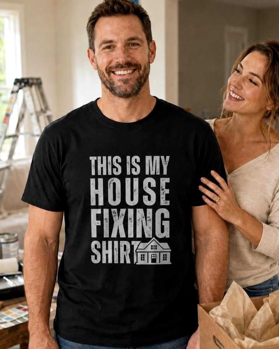 A black short-sleeved t-shirt featuring the white distressed text "THIS IS MY HOUSE FIXING SHIRT" centered on the chest, accompanied by a small graphic illustration of a house with a gabled roof and windows.