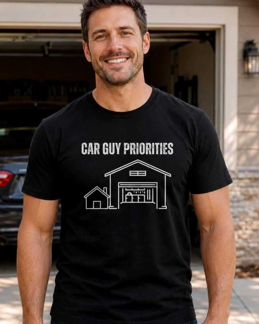 Smiling man wearing a black “Car Guy Priorities” graphic T-shirt while standing in front of his home garage with a car inside.