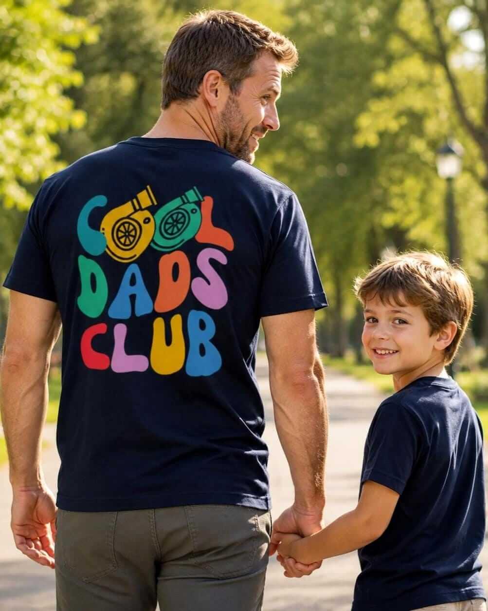 Father in a navy blue Cool Dads Club back-print T-shirt walks hand in hand with his smiling son along a sunny park path.
