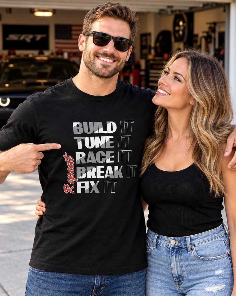 Smiling car enthusiast wearing a black “Build It, Tune It, Race It, Break It, Fix It, Repeat” graphic T-shirt while embracing his wife, with a modified muscle car blurred inside their garage.
