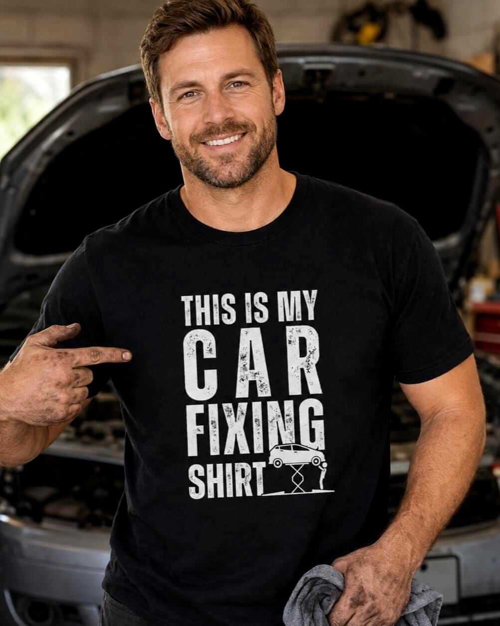 Smiling man wearing a black “This Is My Car Fixing Shirt” T-shirt in a garage, pointing to the white distressed design with greasy hands while a car with its hood open is blurred behind him.