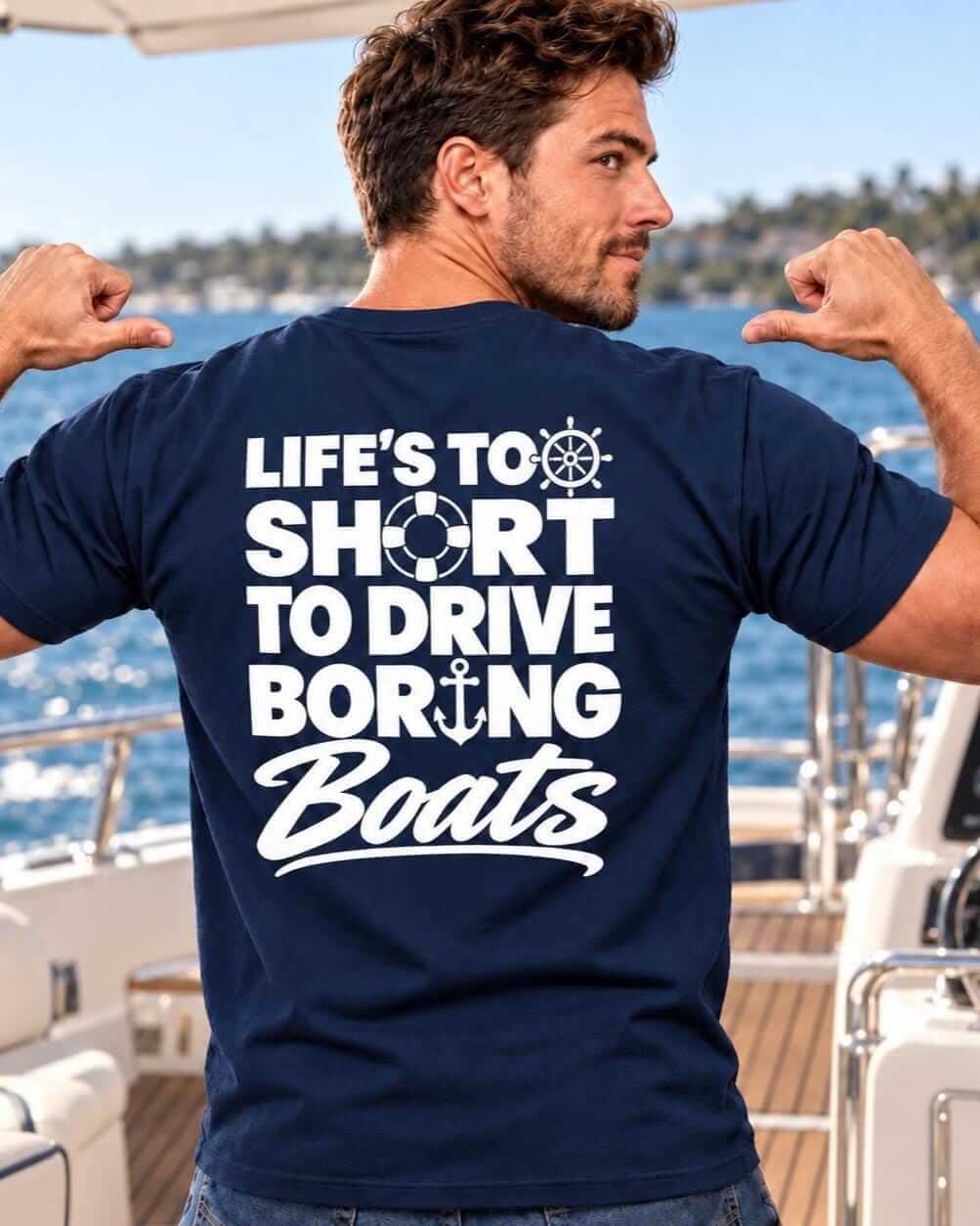 Smiling man wearing a navy blue “Life’s Too Short to Drive Boring Boats” T-shirt, pointing to the white back design while standing on a motorboat with blue water behind him.