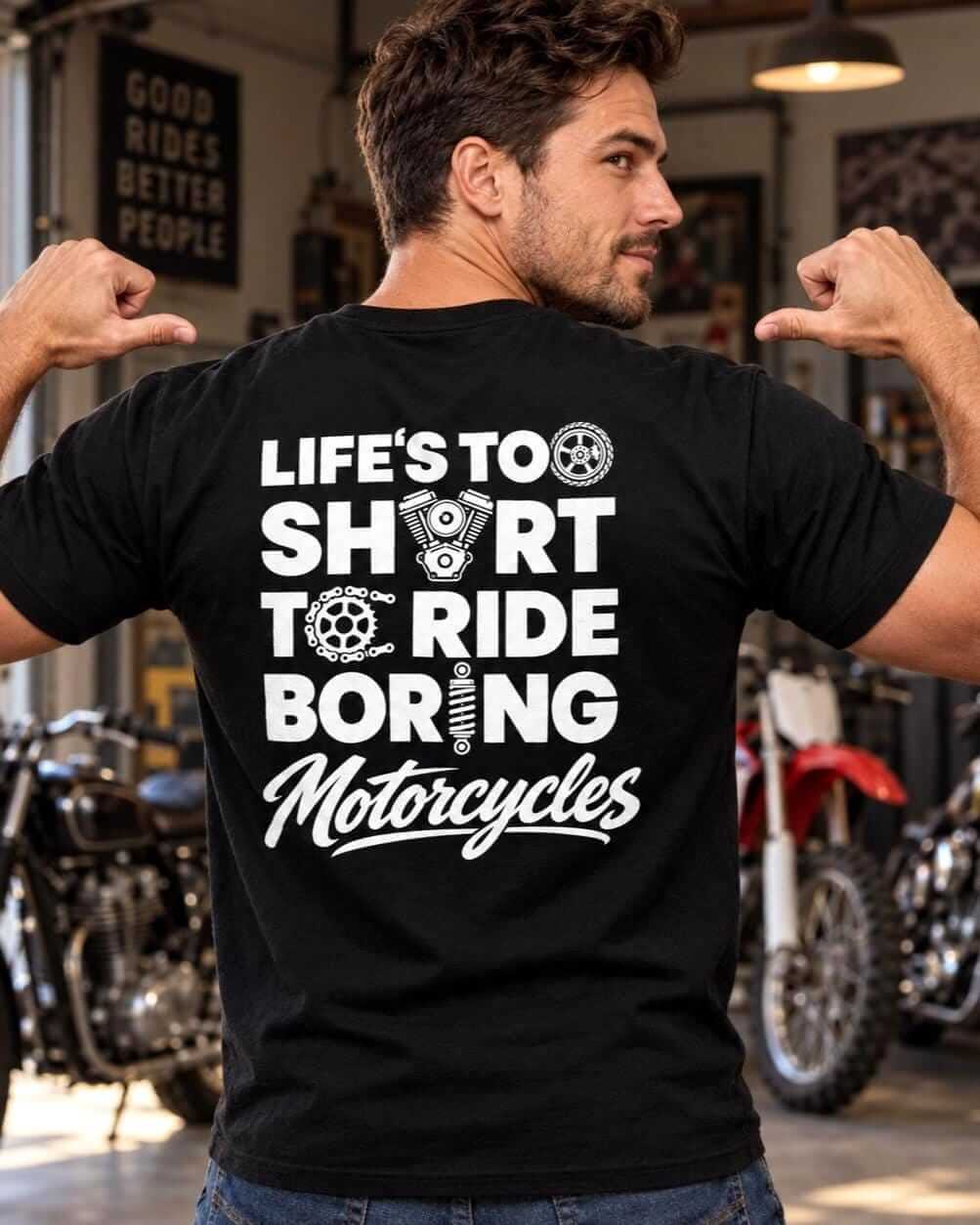 Man wearing a black “Life’s Too Short to Ride Boring Motorcycles” T-shirt, pointing to the back design in a garage with classic, dirt and chopper motorcycles.