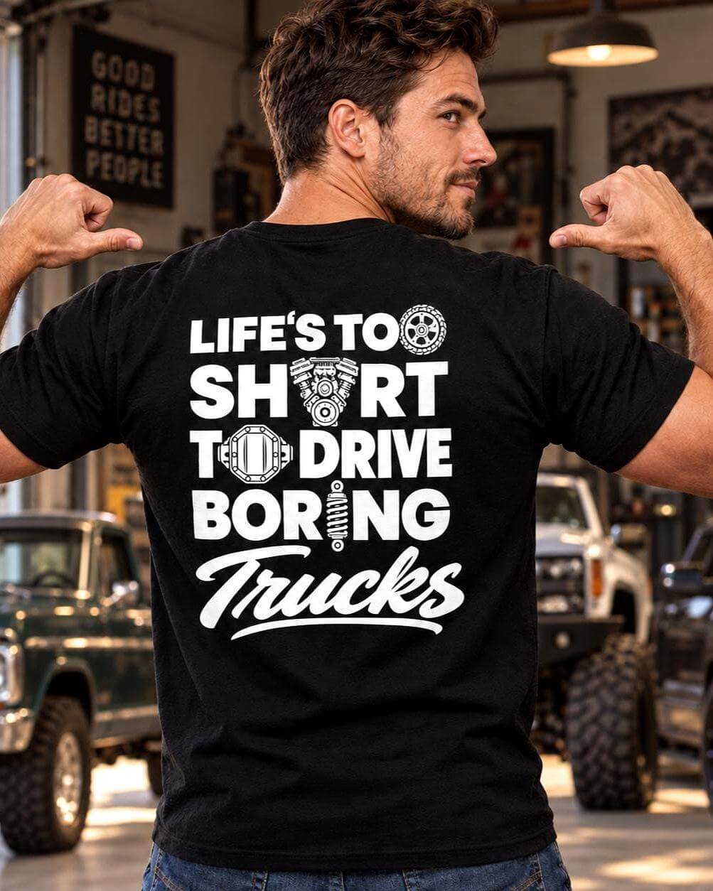 Athletic man wearing a black T-shirt with a white Life’s Too Short to Drive Boring Trucks back design, pointing at the print in a garage with three custom pickup trucks.