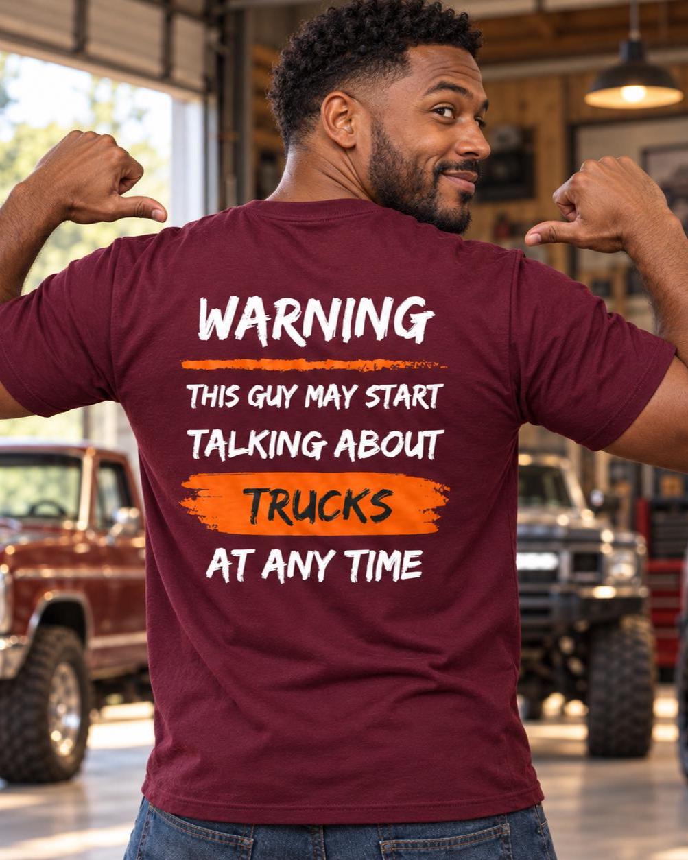 Smiling man shown from behind in a maroon T-shirt, pointing to the “Warning: This Guy May Start Talking About Trucks at Any Time” back design in a warm truck garage setting.