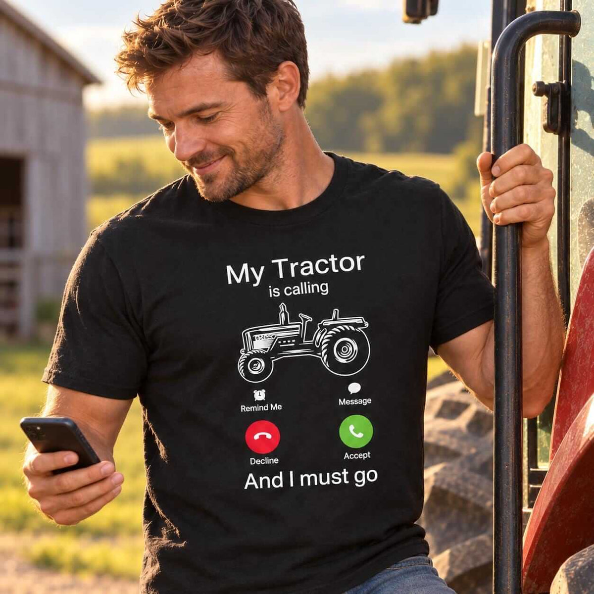 Man standing on a tractor step, looking at his phone and wearing a black “My Tractor is calling – And I must go” T-shirt with a tractor graphic and call icons, beside a barn in golden afternoon light.
