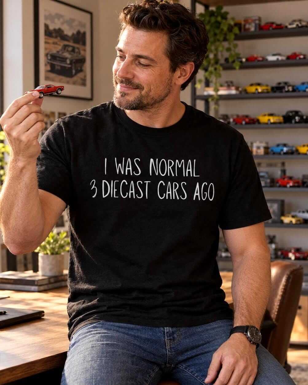 Smiling man wearing a black “I Was Normal 3 Diecast Cars Ago” T-shirt while holding a small model car in a home office with diecast cars displayed on shelves.