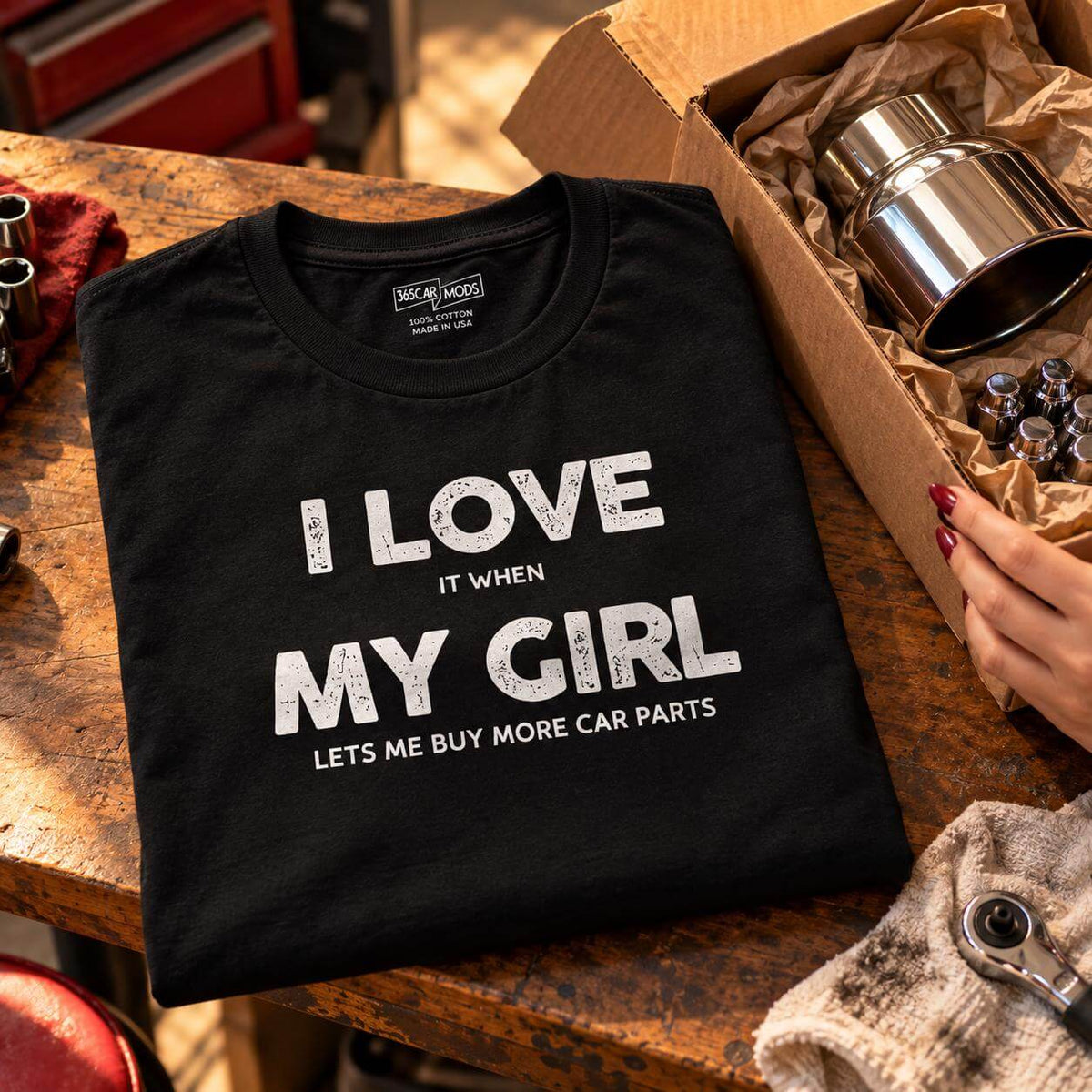 Folded black I Love My Girl car parts shirt next to an open box of new car parts and a woman's hand.