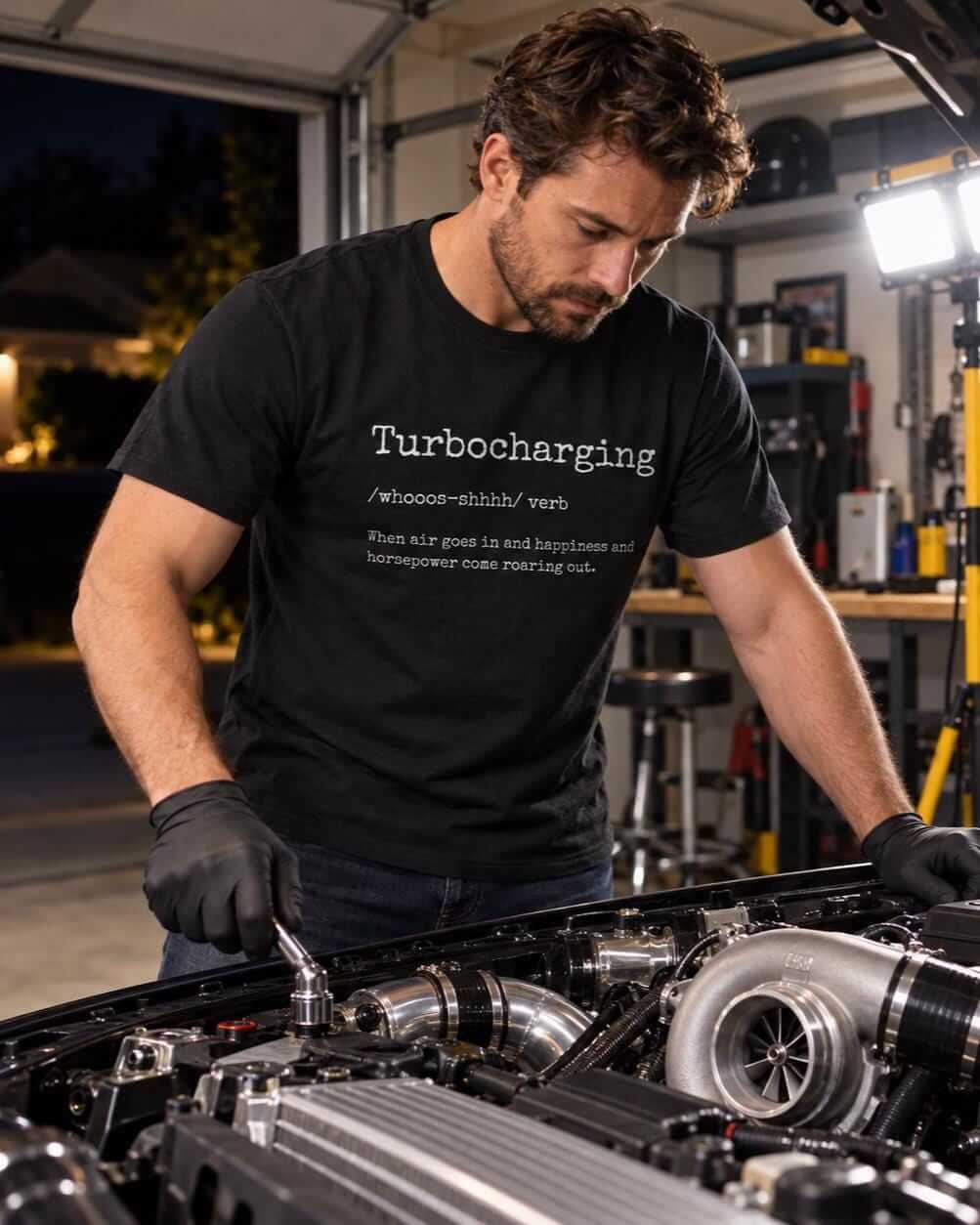 Man in a black turbocharging definition shirt leaning over an engine bay with a turbocharger in a home garage at night