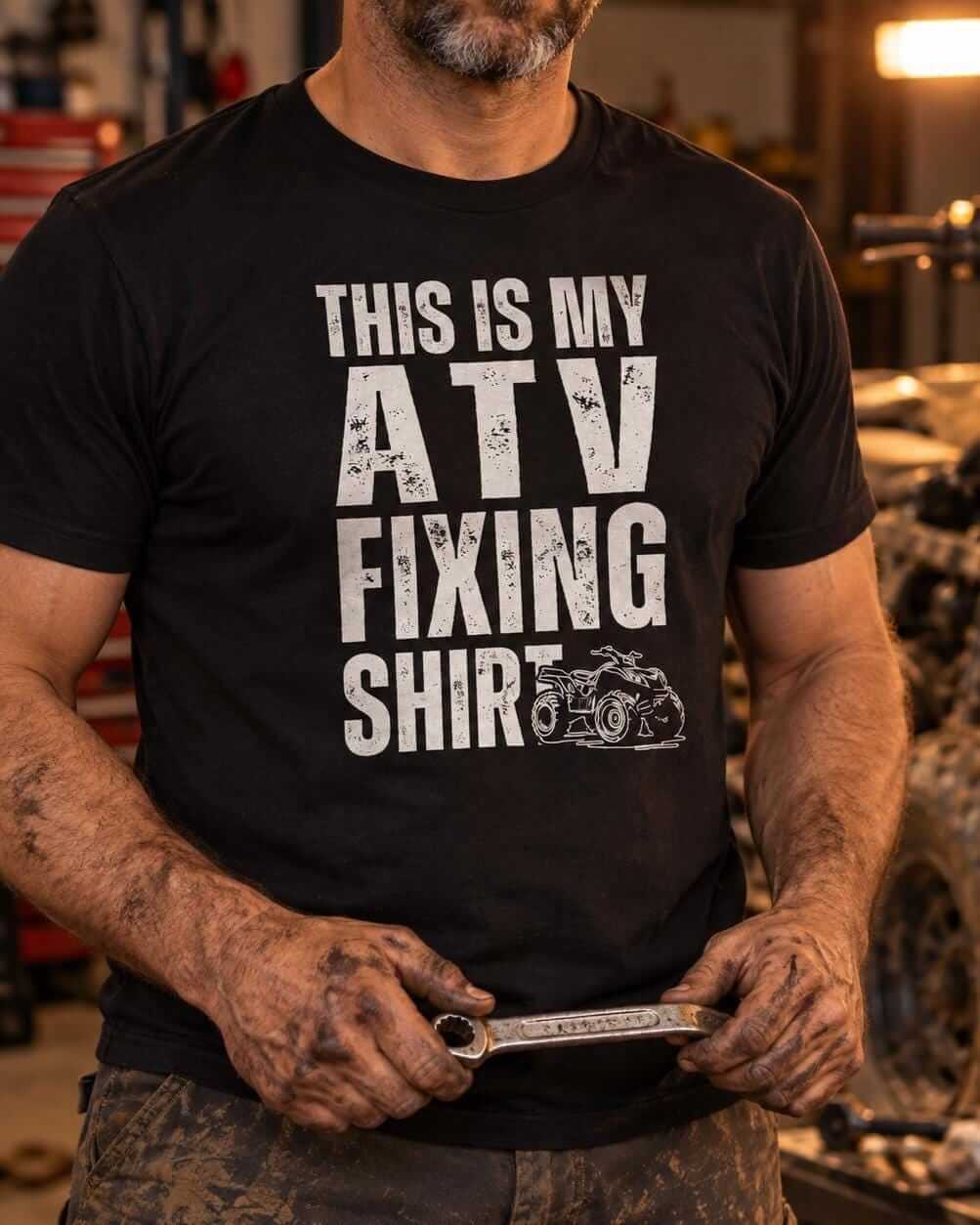 Close-up of a man with greasy hands wearing a black This Is My ATV Fixing Shirt in a garage with a quad