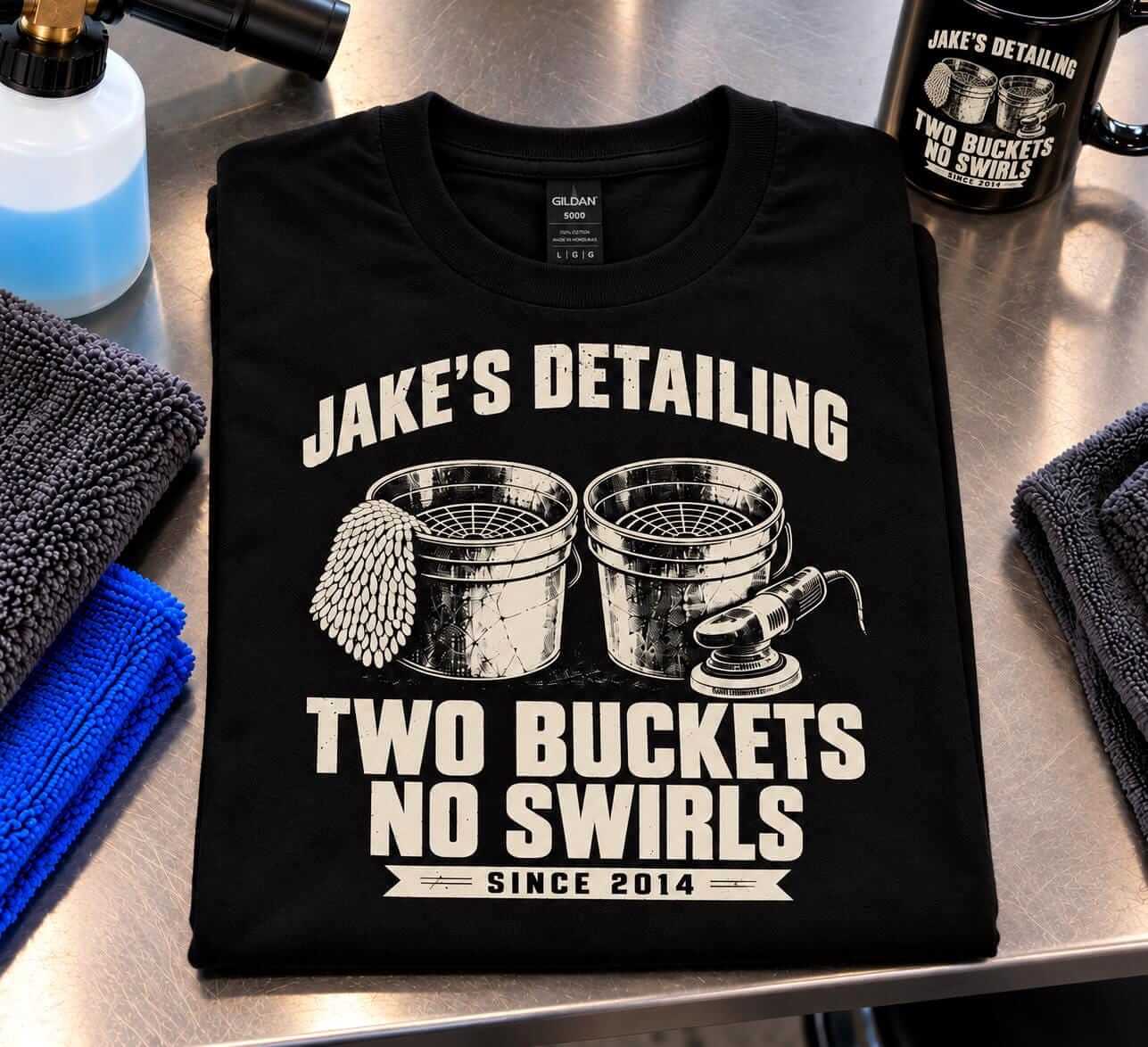 Black Two Buckets No Swirls personalized detailing t-shirt folded on a garage counter next to microfiber towels