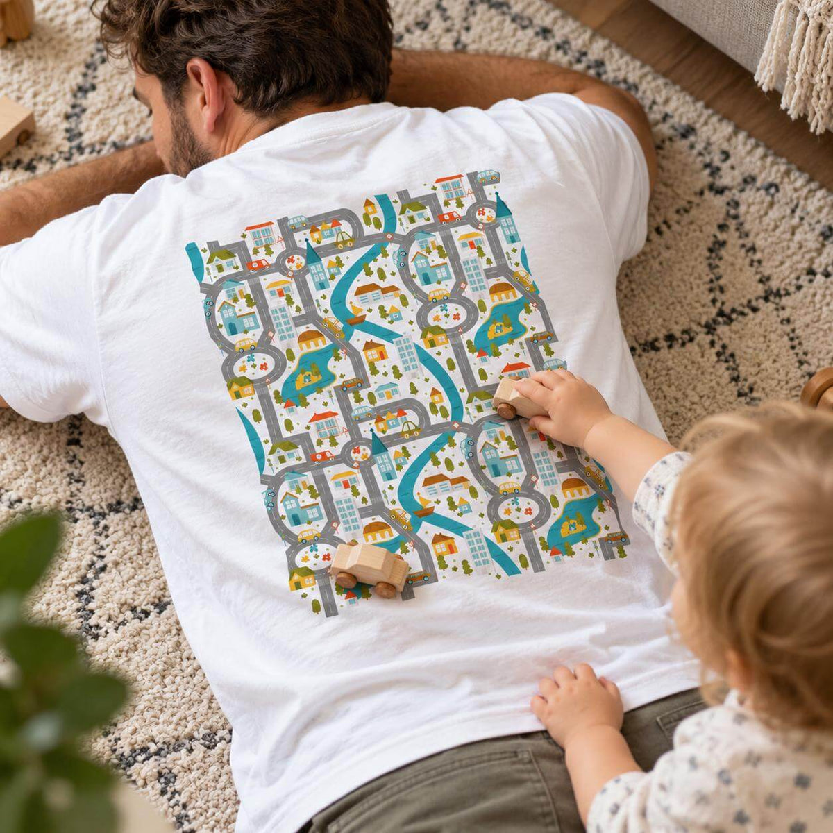 Dad lying on a rug in a white t-shirt with a road play mat printed on the back while a toddler drives a toy car on it