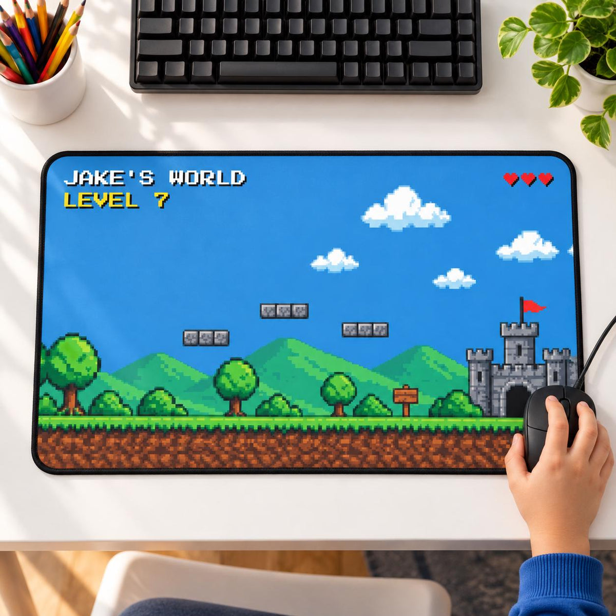 Personalized pixel art gaming desk mat with JAKE'S WORLD and LEVEL 7, hills and a castle, with a child's hand on the mouse on a white desk