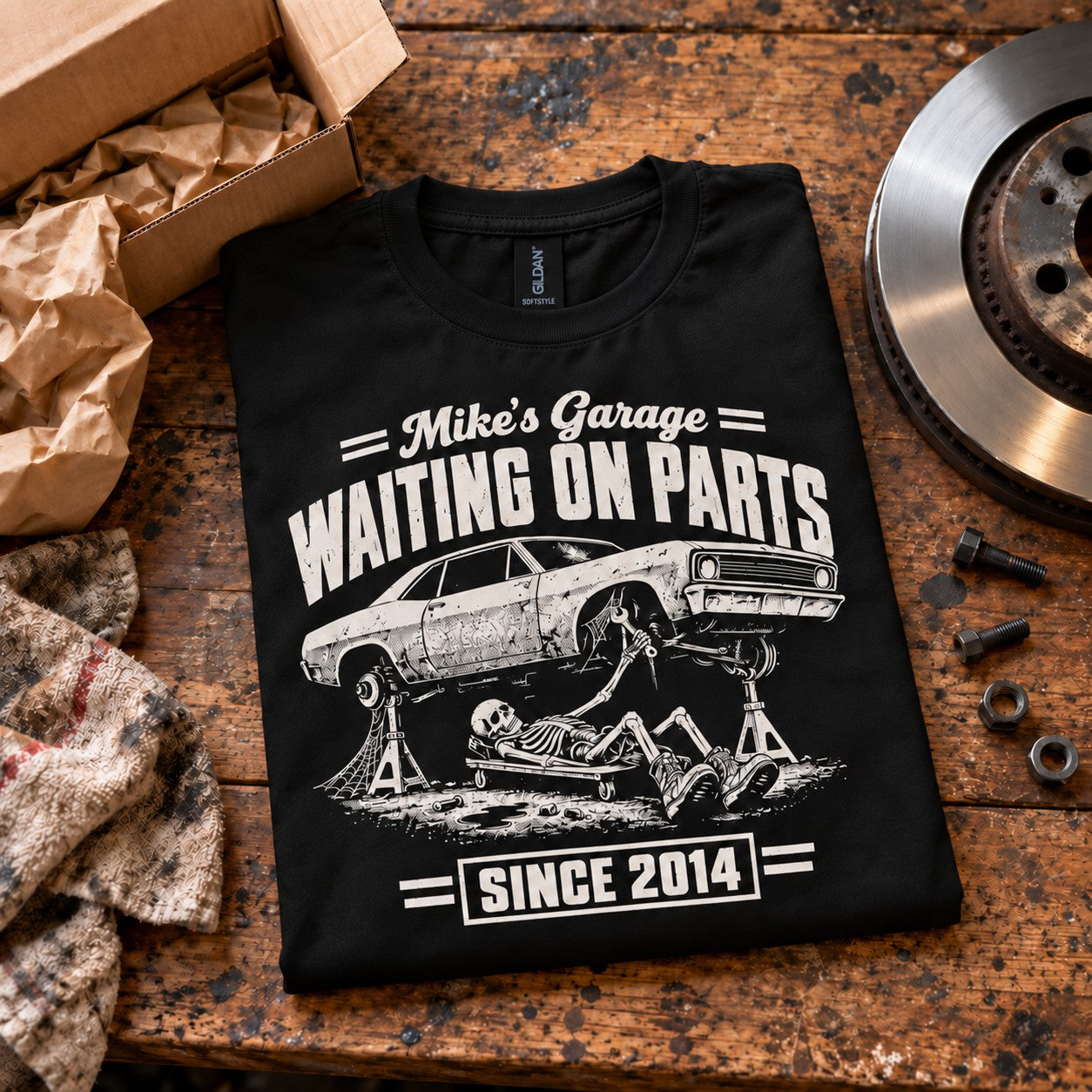 Black t-shirt with WAITING ON PARTS, a skeleton mechanic on a creeper under a project car on jack stands, Mike’s Garage on top and Since 2014 below